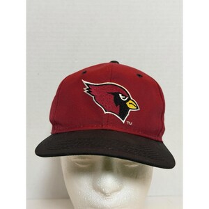 Könnte beinhalten: Eine kastanienbraune Baseballkappe mit schwarzem Schirm und einem gestickten Kardinalvogel-Logo auf der Vorderseite. Der Vogel ist im Profil mit gelbem Schnabel und schwarzem Auge dargestellt. Die Buchstaben TM sind klein und weiß darunter gestickt.