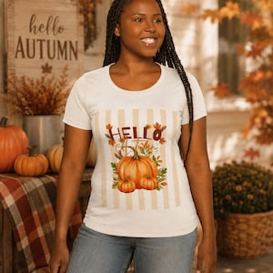 Könnte beinhalten: Ein weißes Kurzarm-T-Shirt mit einer zentralen Grafik aus drei orangefarbenen Kürbissen, umgeben von Herbstblättern und dem Schriftzug "HELLO Autumn" in brauner und goldener Schrift, vor einem Hintergrund mit beigen Streifen.