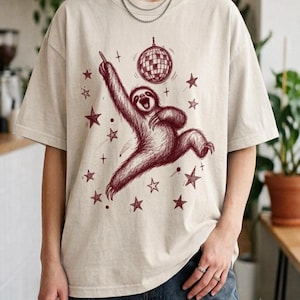 Könnte beinhalten: Ein beiges T-Shirt mit kurzen Ärmeln und einer dunkelroten grafischen Illustration eines Faultiers, das mit einem erhobenen Arm zu einer Discokugel tanzt, umgeben von verschiedenen Sternen. Das Design ist im Skizzenstil auf der Vorderseite.