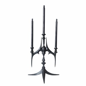 May include: A matte black metal candelabra featuring a central pillar and two side arms, each holding a slender black taper candle. The structure has a pointed, angular base with three feet and decorative, sharp-edged accents at the joints.