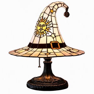 May include: A stained glass table lamp shaped like a pointed witch hat. The shade features a cream background with a yellow sun face, scattered stars, and a dark band with a buckle. It sits on a dark, ornate metal base with a pull chain switch.