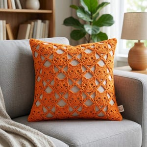May include: A square throw pillow featuring a bright orange crocheted cover with a repeating cat face pattern. The openwork design reveals a solid light-colored fabric underneath. A small white tag with a cat icon is attached to the side.