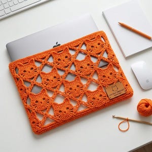 May include: An orange crochet laptop sleeve featuring a repeating pattern of cat faces. A rectangular brown leather tag is attached to the lower right corner with the text "CAT FACE CROCHET - Handmade -" and a small cat icon engraved on it.