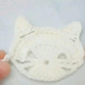 May include: A white crocheted cat face applique featuring pointed ears, openwork eye details, and a small circular nose area. The piece is crafted from white yarn with a visible loose thread extending from the left side of the cat head.