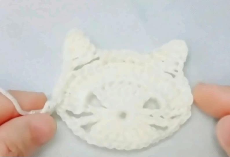 May include: A white crocheted cat face applique featuring pointed ears, openwork eye details, and a small circular nose area. The piece is crafted from white yarn with a visible loose thread extending from the left side of the cat head.
