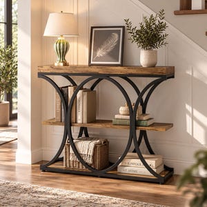 Peut inclure: Une console avec un plateau et deux étagères en bois brun veiné, soutenus par une structure en métal noir avec des accents circulaires latéraux. La table supporte une lampe, un cadre avec une plume, une plante, des livres et un panier en osier.
