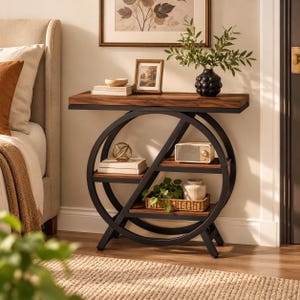 Peut inclure: Une table console avec un plateau en bois brun foncé et deux étagères inférieures soutenues par un cadre circulaire en métal noir. La table porte un vase noir texturé avec de la verdure, un petit cadre photo, des livres, une radio et un panier.