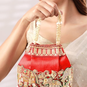 May include: A red satin handbag featuring gold embroidered elephant motifs and decorative sequins. The bag has a structured base and a handle made of multiple strands of white faux pearls, accented with larger pearl beads at the connection points.