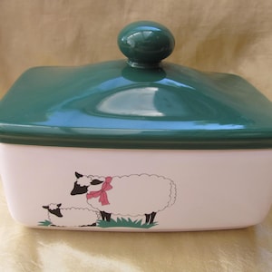 Könnte beinhalten: Eine rechteckige Butterdose aus Keramik mit einem glänzenden, dunkelgrünen Deckel und einem runden Knauf. Der untere Teil ist cremeweiß und mit einer grafischen Illustration eines großen Schafes mit rosa Schleife und eines Lamms auf Gras verziert.