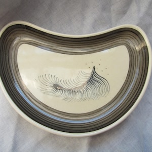 May include: A crescent-shaped ceramic dish featuring a cream-colored center with a black hand-painted feather illustration and small dots. The rim is decorated with concentric dark gray and black circular bands, creating a layered effect.