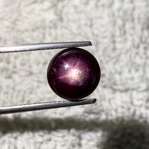 Peut inclure: Une pierre précieuse cabochon ronde d'un violet profond présentant un effet d'étoile à six branches distinct centré sur sa surface. La pierre est maintenue fermement entre les deux pointes métalliques d'une pince, avec une finition lisse et polie.