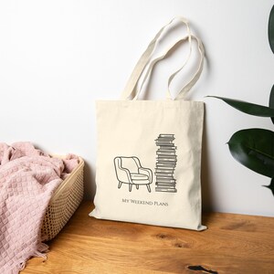 May include: A cream-colored canvas tote bag featuring a black line drawing of an armchair and a tall stack of books. Below the illustration, the text reads "MY WEEKEND PLANS" in a simple, clean font. The bag has two long shoulder straps.