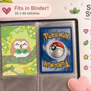 May include: A black binder containing a cross stitch pattern of a brown and white owl-like creature on a green background, placed next to a blue Pokémon trading card. Text includes "Fits in Binder! 32 x 46 stitches" and "Shiny Stitch."
