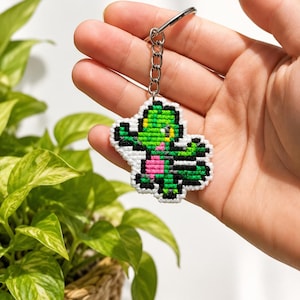 Op de afbeelding: Een in kruissteek geborduurde sleutelhanger met een gepixelde groene hagedisachtige figuur met een roze borst en zwarte accenten, bevestigd aan een zilverkleurige metalen ring en ketting. Het ontwerp is omlijnd met wit garen.