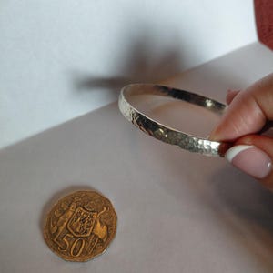 May include: A silver-coloured bangle bracelet with a hammered, textured surface. The bracelet is positioned next to an Australian fifty-cent coin, which features a coat of arms with two kangaroos and the number 50, providing a scale reference.