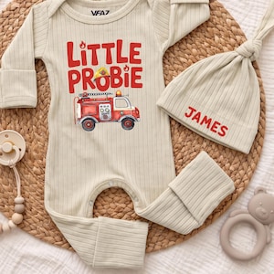 May include: A beige ribbed long-sleeve baby bodysuit featuring a red fire engine graphic and the text Little Probie, paired with a matching ribbed beanie embroidered with the name James, a beige dummy clip with wooden beads, and a bear teether.