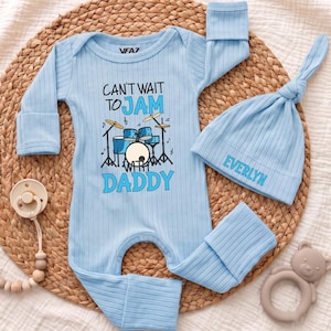 May include: A light blue ribbed long-sleeve baby bodysuit and matching knotted hat. The bodysuit features a graphic of a drum set with the text "CAN'T WAIT TO JAM WITH DADDY". The hat is embroidered with the name "EVERLYN". Includes a pacifier clip.