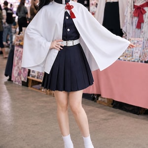 May include: A dark navy blue pleated skirt and matching high-collar jacket with silver buttons, worn under a white cape featuring a red decorative knot. The outfit includes a white belt, white socks, and black thong sandals with a butterfly hair clip.