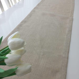 May include: A long, rectangular table runner made of light brown, woven burlap fabric with a visible textured weave. The runner is positioned lengthwise, accompanied by a small bouquet of white artificial tulips with green stems.
