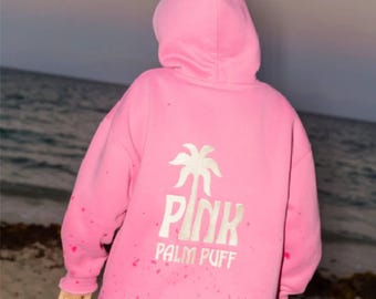 May include: A bright pink hooded sweatshirt featuring a white graphic on the back. The graphic displays a stylized palm tree above the text "PINK" and "PALM PUFF" in a bold, sans-serif font. The fabric has a subtle, speckled texture throughout.