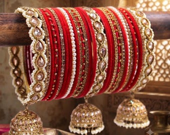 May include: A set of red velvet bangles featuring gold-tone metal accents, rows of small white pearls, and clear crystal embellishments. Three gold-tone bell-shaped jhumka charms with pearl trim hang from the bangles, creating a layered design.