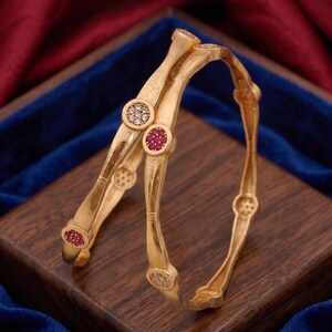 Op de afbeelding: Twee goudkleurige armbanden met een textuur die doet denken aan bamboe. Elke armband is versierd met afwisselende ronde zettingen die ofwel kleine, heldere witte steentjes of levendige rode steentjes bevatten op het buitenoppervlak.