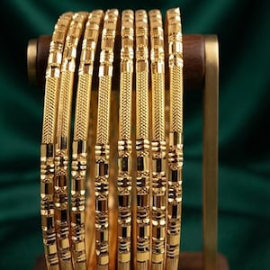 Op de afbeelding: Een set van zeven gouden armbanden met afwisselend getextureerde en gepolijste segmenten. De armbanden zijn verticaal op een houten standaard geplaatst. De tekst GOLD SHINING is bovenaan de afbeelding zichtbaar. Elke armband toont een herhalend patroon van kruislings gearceerde etsen en gladde, reflecterende gouden oppervlakken.