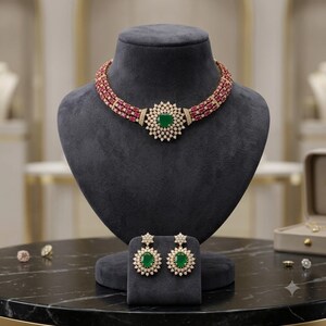 Puede incluir: Un conjunto de collar y pendientes a juego con tres filas de piedras rojas rectangulares. El collar tiene un colgante circular central con una piedra verde rodeada de piedras blancas. Los pendientes reflejan este diseño circular.