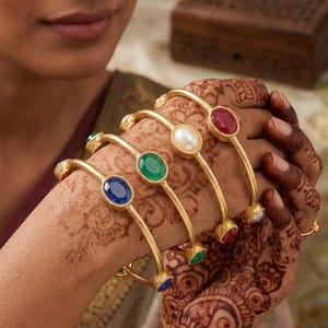 May include: A set of four gold-toned bangles, each featuring a single oval-shaped gemstone. The stones include a red ruby, a white pearl, a green emerald, and a blue sapphire. Each bangle has a textured gold finish and a simple metal band design.