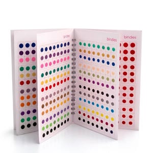 May include: A spiral-bound booklet containing multiple pages of small, circular adhesive stickers in a wide variety of colours. Each page features rows of dots arranged in a grid pattern. The word bindies is printed in small pink text on the pages.