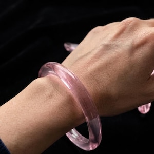 May include: A translucent, light pink bangle bracelet with a smooth, rounded surface. The bracelet is circular in shape and features a clear, glass-like appearance, reflecting light across its polished, curved exterior.