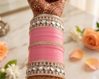 May include: A stack of bangles featuring solid pink bands interspersed with two decorative bands containing clear, faceted stones and small red and green accents. A large, circular, multi-stone ring is worn on the finger above the bangles.
