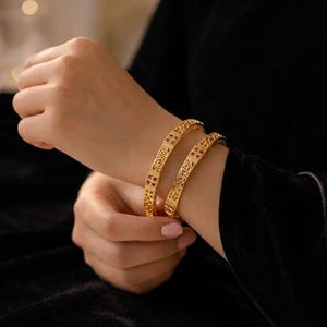 Könnte beinhalten: Zwei goldfarbene Armreifen mit einem komplizierten durchbrochenen Filigranmuster und kleinen, quadratisch geschliffenen roten Steinen, die in regelmäßigen Abständen entlang des Umfangs jedes kreisförmigen Armbands eingearbeitet sind.