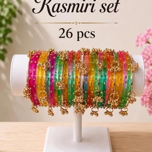 May include: A Kasmiri set of 26 pieces consisting of translucent, multi-coloured bangles in shades of pink, yellow, green, and orange. Each bangle features small, gold-toned metallic charms hanging from the outer edge, arranged on a white stand.