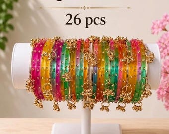 May include: A set of 26 Kasmiri bangles in translucent pink, yellow, and green colors. Each bangle features small gold-toned metal charms attached to the outer rim. The bangles are displayed on a white cylindrical stand with the text Kasmiri set 26 pcs.