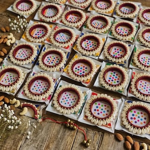 May include: A collection of circular bangles featuring a deep maroon fabric base adorned with a border of small white cowrie shells. Each bangle is individually packaged in a clear plastic sleeve with a colorful polka dot card insert.