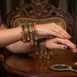 May include: A set of multi-colored translucent glass bangles featuring a gold-toned metal frame. Each bangle is adorned with small, dangling gold-toned bell charms. The bangles are arranged in a stack, showcasing vibrant red, green, and blue hues.