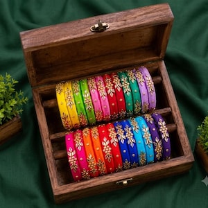 Pode incluir: Uma coleção de pulseiras multicoloridas dispostas em duas fileiras dentro de uma caixa de madeira. Cada pulseira apresenta um acabamento de tecido liso e vibrante, adornado com um padrão floral dourado central com pequenas pedras refletoras.