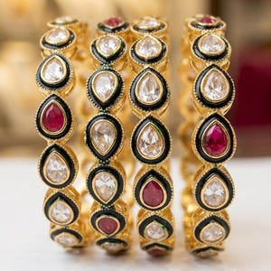 May include: A set of four gold bangles featuring a repeating teardrop pattern. Each teardrop shape is outlined in black enamel and contains either a clear, faceted stone or a deep red, faceted stone, creating a vibrant and detailed jewelry piece.