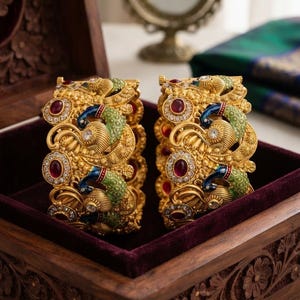 Op de afbeelding: Een paar goudkleurige armbanden met gedetailleerde pauwenmotieven en groene en blauwe emaille details. De armbanden zijn versierd met ronde rode stenen en kleine heldere kristallen die in een gestructureerd metalen oppervlak zijn gezet.