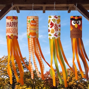 May include: Four cylindrical wind socks featuring Thanksgiving themes. The first displays autumn leaves and the text HAPPY THANKSGIVING. The second shows stacked pumpkins with the words Thankful, Grateful, Blessed. The third has fall leaves, and the fourth depicts a turkey.