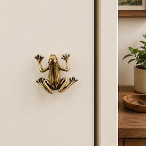 May include: A brass-toned metal cabinet knob shaped like a frog with its limbs extended. The frog figure has a textured, metallic finish and is mounted onto a flat, light-colored surface, serving as a functional handle for a door or drawer.