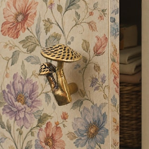 May include: A brass-toned metal cabinet knob shaped like two mushrooms with textured caps, mounted on a light-colored cabinet door featuring a printed pattern of large pink, purple, and blue flowers with green leaves and thin brown stems.