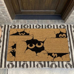 May include: A rectangular tan coir doormat featuring a black graphic print of four distinct cracks in the surface, each revealing a pair of wide, circular eyes peering out from the dark voids. The design creates a layered, peek-a-boo effect.