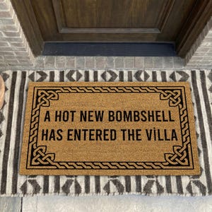 May include: A rectangular tan coir doormat featuring a black border with a geometric knot pattern and the text A HOT NEW BOMBSHELL HAS ENTERED THE VILLA printed in black capital letters in the center of the mat.