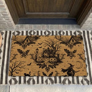 May include: A rectangular tan coir doormat featuring a black ink illustration of a haunted house, two jack-o'-lanterns, a flying witch, bats, a crescent moon, and a black cat, layered over a larger rectangular rug with a brown and white geometric pattern.