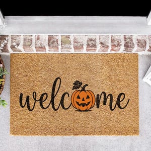 May include: A rectangular tan coir doormat featuring the word welcome in black cursive script. The letter o is replaced by a graphic of an orange jack-o'-lantern pumpkin with a black stem and leaf, centered on the mat's textured natural fiber surface.