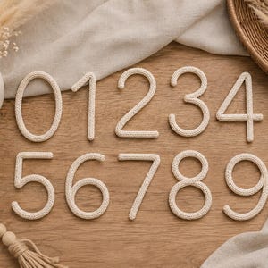 May include: A set of ten off-white knitted numbers from zero to nine, each featuring a textured yarn surface. The numbers are arranged in two horizontal rows, with zero through four in the top row and five through nine in the bottom row.