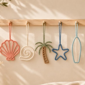 May include: A light wood wall rack with five wooden pegs, each holding a hanging crochet ornament. The ornaments include a coral seashell, a cream snail shell, a green palm tree, a blue starfish, and a light blue surfboard shape.