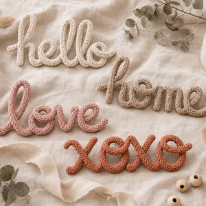 以下が含まれることがあります： クリーム色の「hello」、ダスティピンクの「love」、ベージュの「home」という文字が編み込まれた3つの壁掛けサインと、テラコッタ色の「xoxo」と書かれた別の編み込みサイン。これらの装飾的な筆記体の文字は毛糸で作られています。